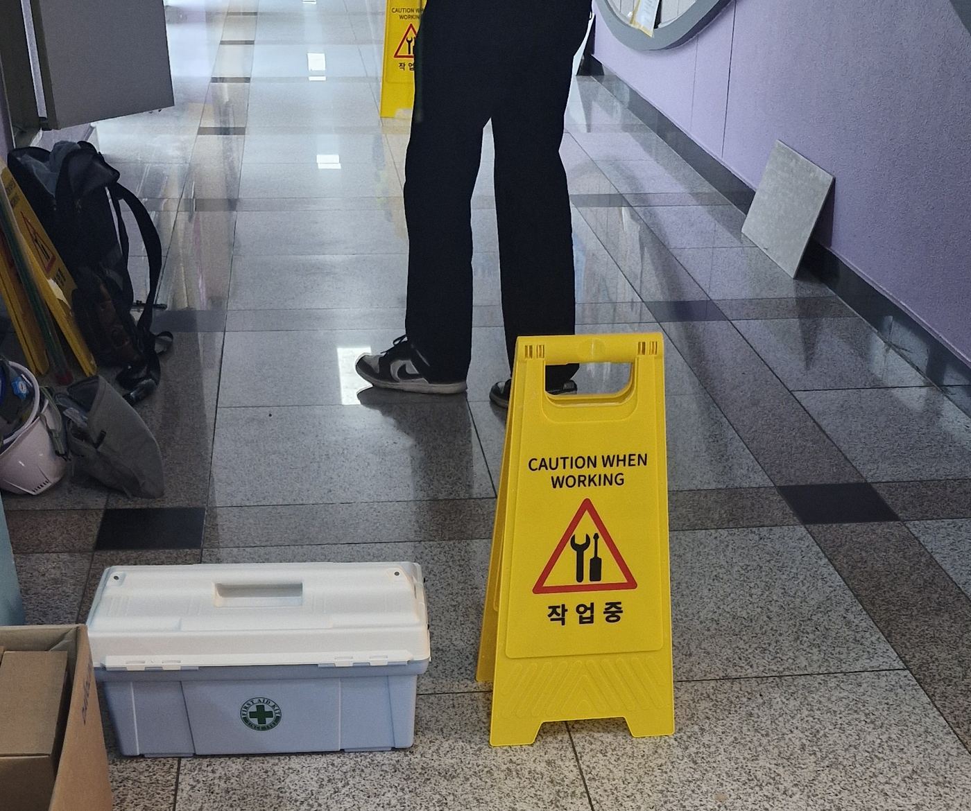 학교 복도 작업 현장, 바닥에 놓인 안전 표지판(CAUTION WHEN WORKING, 작업중)과 구급함, 작업자 하반신